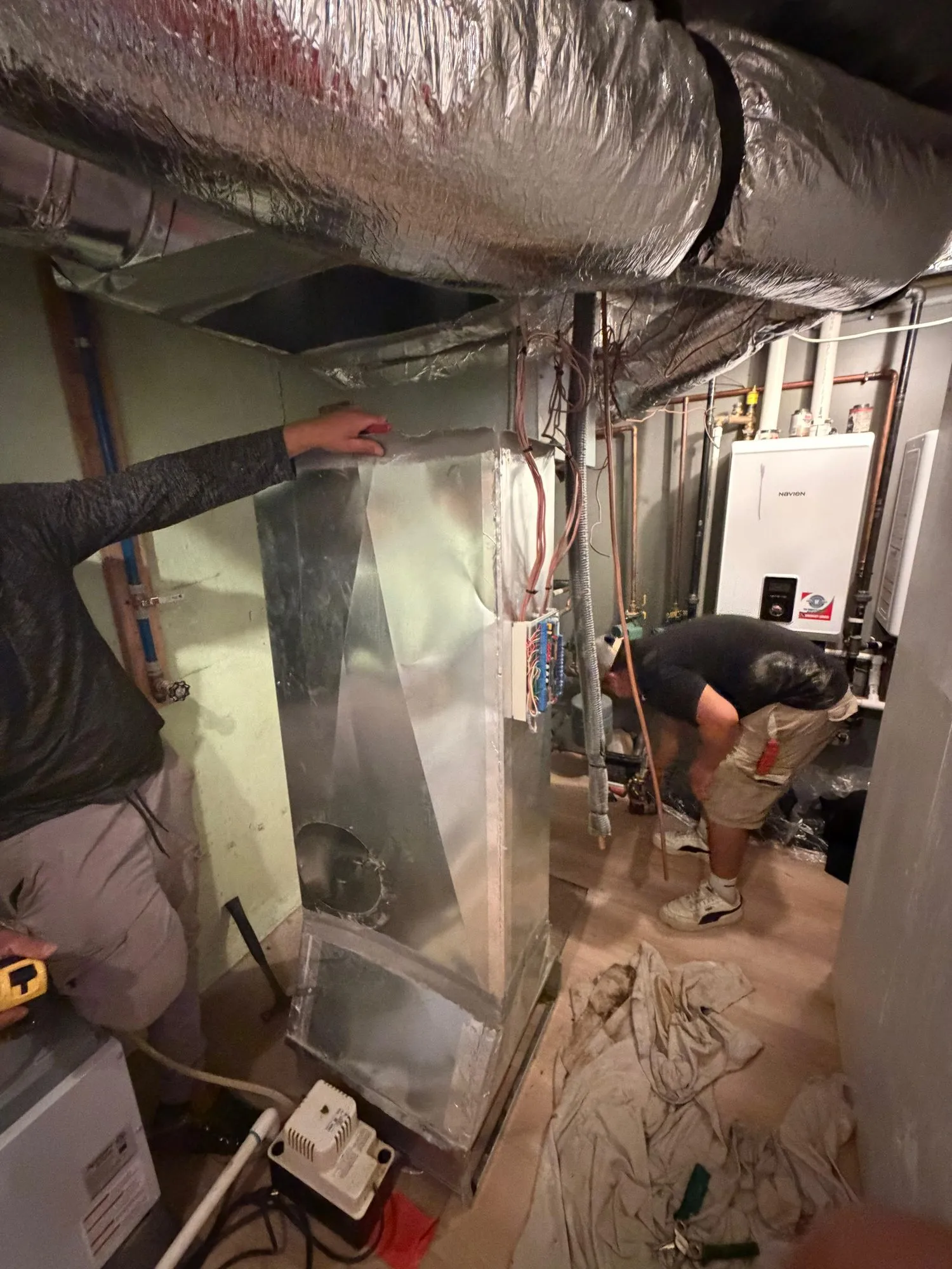 Two technicians fitting a sheet-metal plenum and flex ductwork for an HVAC system