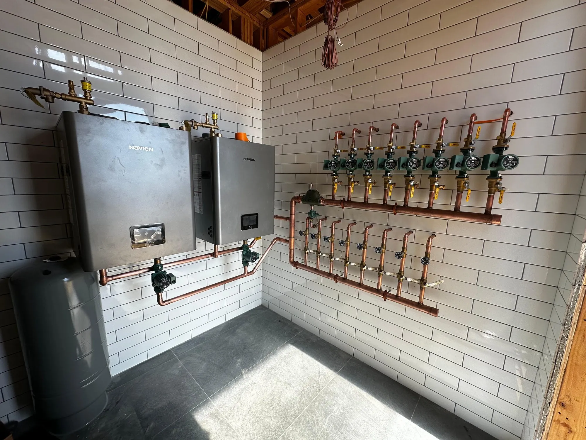 Tiled mechanical room with two wall-hung Navien units, an expansion tank and a copper radiant heating zone manifold